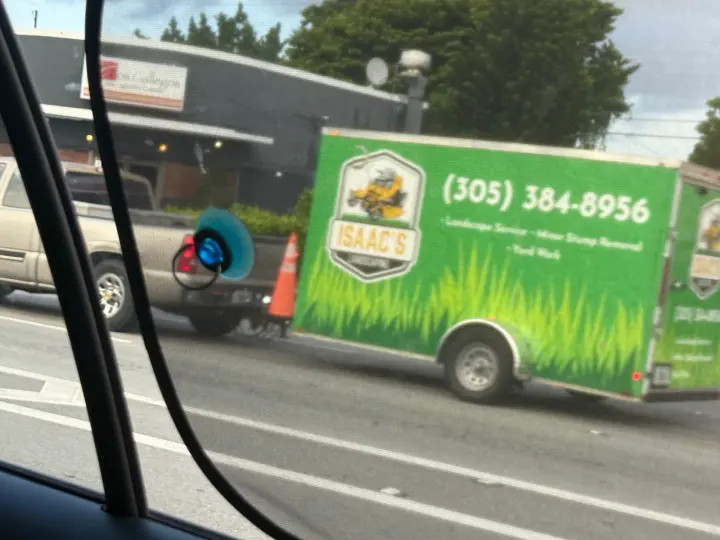 Green Isaac's lawn-service trailer displaying a mower logo and phone number 305-384-8956